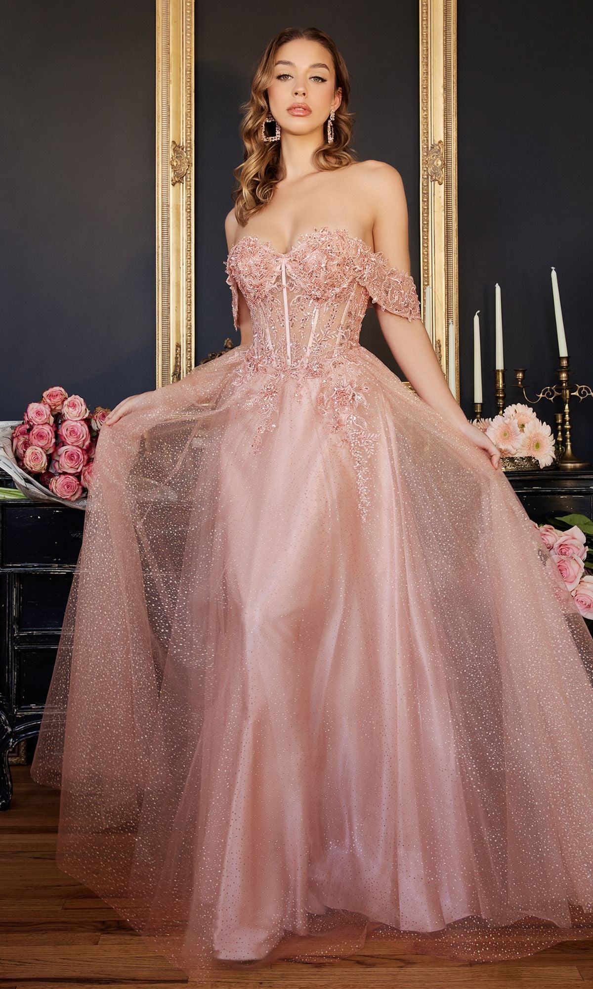 A woman stands indoors wearing the Off-Shoulder Long Glitter A-Line Prom Dress CD0198 in blush pink, with embroidered lace and floral details. She holds her gown's sides; flowers and candelabras decorate the background.